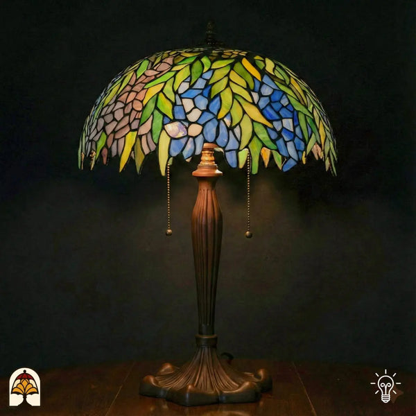 Verlichte grote Tiffany tafellamp met brede koepelvormige kap vol groene bladeren, blauwe en roze/lila bloemen en gele accenten, bronzen kandelaarvoet met trekschakelaars, donkere achtergrond