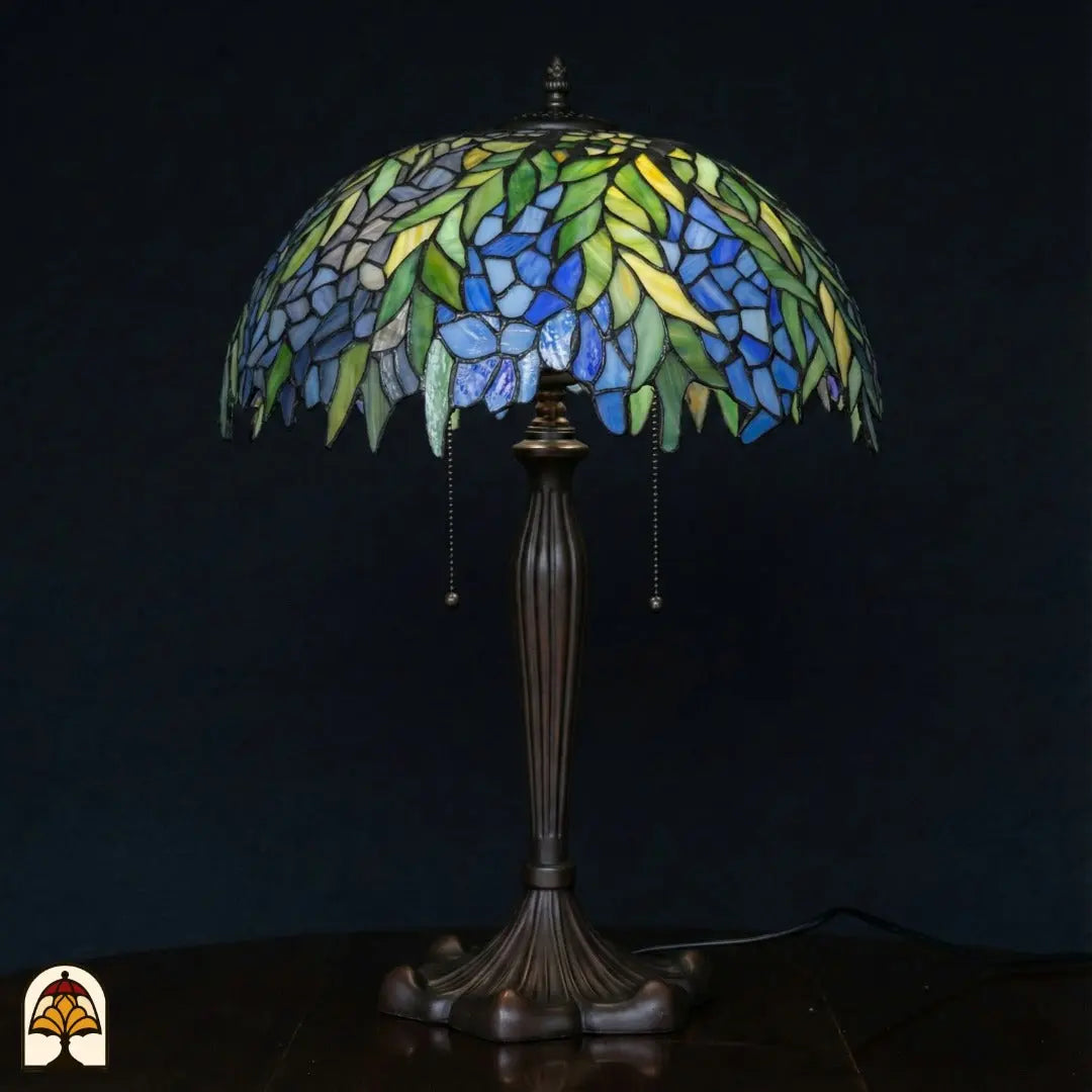 Onverlichte Tiffany tafellamp op donkerblauwe achtergrond — brede koepelvormige kap met groene bladeren en blauwe bloemen, bronzen voet met trekschakelaars