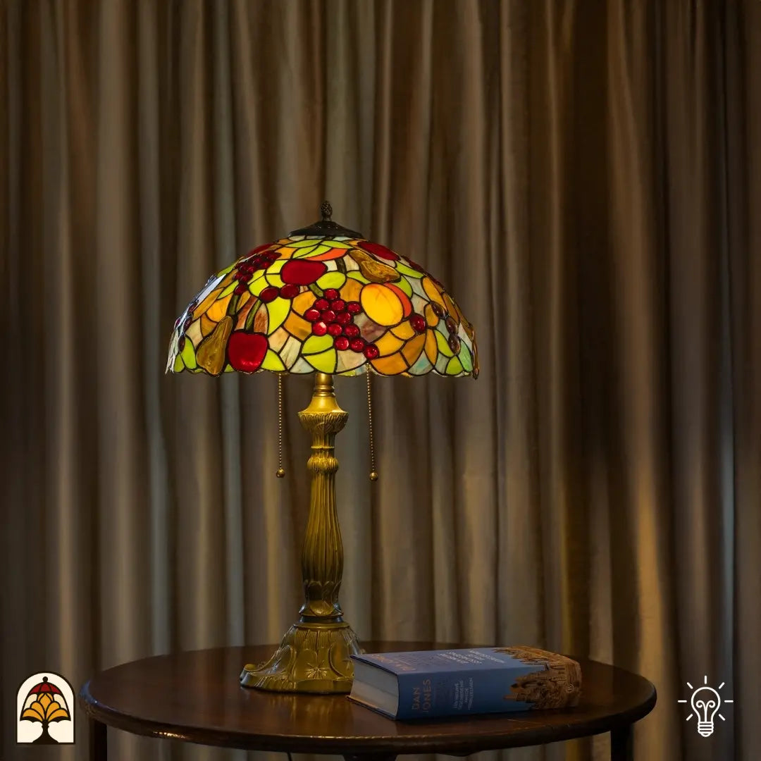 Tiffany tafellamp Frutta Viva verlicht op ronde bijzettafel voor beige gordijnen — boek en decoratiefstuk rechts naast de lamp