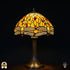 Verlichte Tiffany tafellamp Libellula Aurea ⌀30 op donkere achtergrond, koepelvormige kap met goudgele glas-in-lood panelen, libellenfries onderaan met beige vleugels, rode en groene ovale juwelen, bronzen trompetvoet