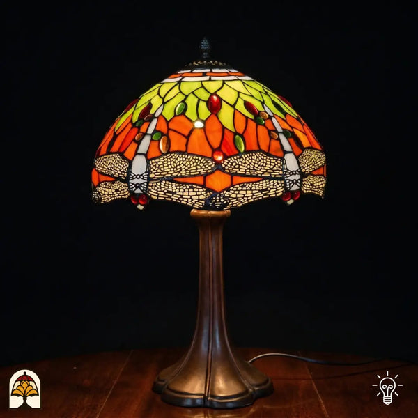 Verlichte Tiffany tafellamp Libellula Azul ⌀30 op donkere achtergrond, koepelvormige kap met oranje glas-in-lood panelen, geel-groene bladmotieven, libellenfries onderaan met beige vleugels, rode en groene ovale juwelen, bronzen trompetvoet