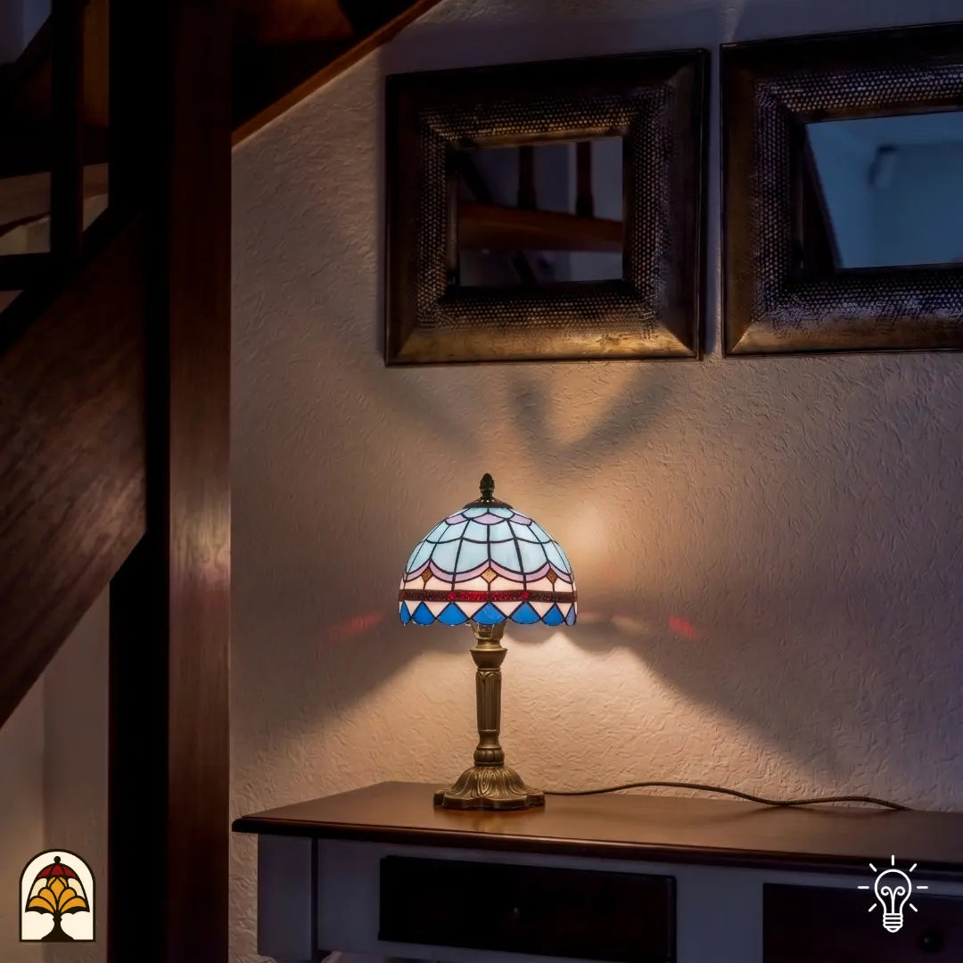 Een kleine, sfeervolle Tiffany tafellamp met een kap in een blauw-wit schubbenpatroon en een donkere sierrand. De lamp staat op een elegante, bronskleurige voet op een houten dressoir. Tegen de achtergrond van een witte muur met decoratieve spiegels en een houten trap verspreidt de lamp een zacht, warm licht dat subtiel op de wand schijnt.