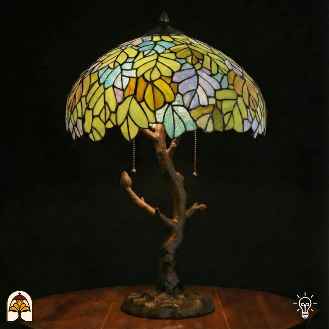 Verlichte Tiffany tafellamp Sylva Verde met brede koepelvormige kap vol geel-groene, geel-oranje en lichtblauwe bladmotieven in glas-in-lood, verweerde bronzen boomstam voet met vertakkingen en trekschakelaars, donkere achtergrond