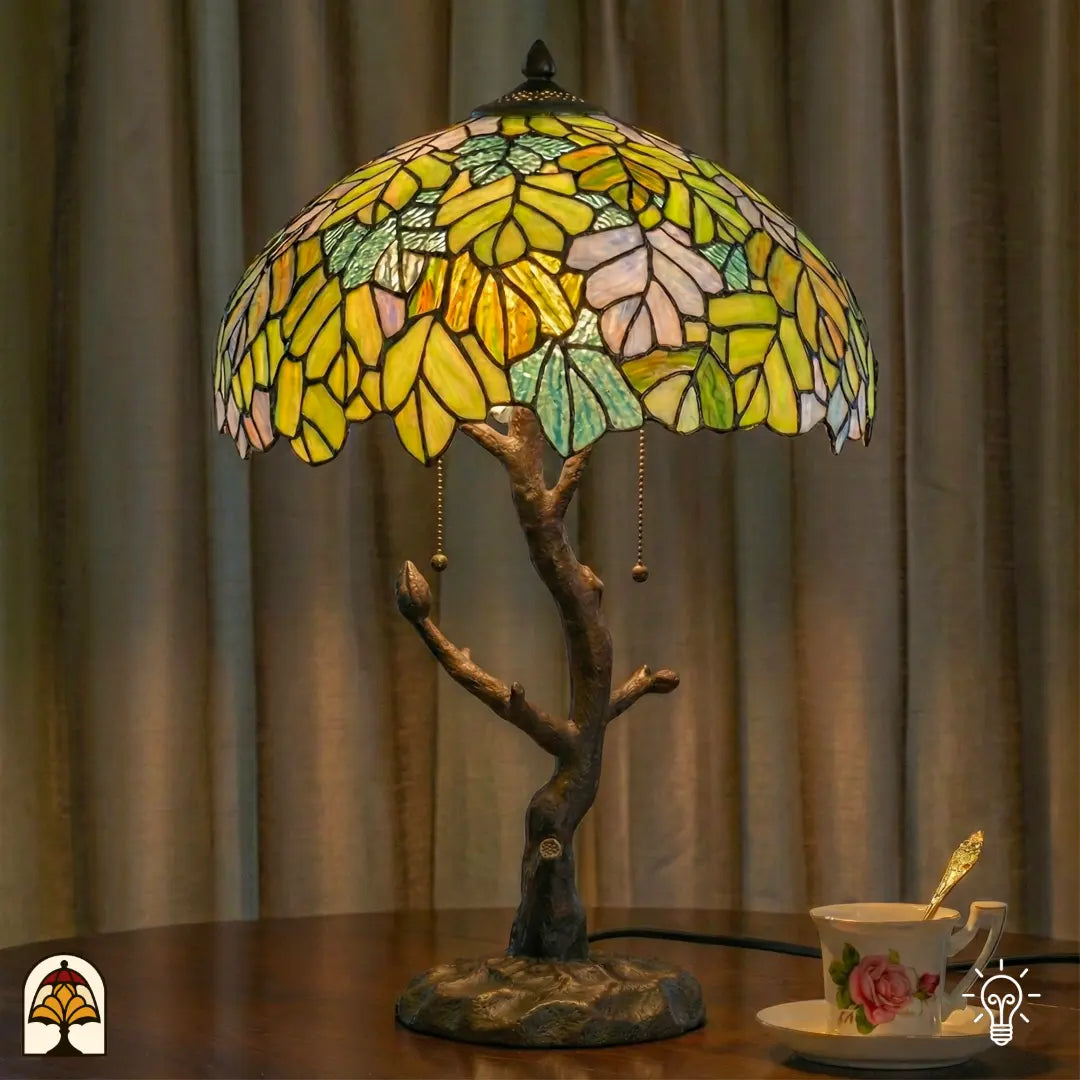 Verlichte Tiffany tafellamp Sylva Verde op houten tafel voor gordijnen, geel-groene en oranje bladmotieven verlicht, theekopje met schotel ernaast