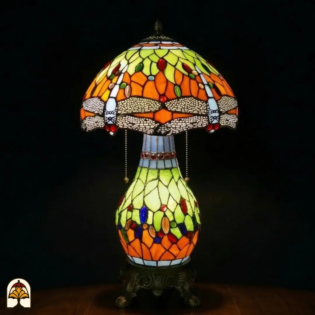 Verlichte Tiffany tafellamp Libellula Lumina ⌀30 op donkere achtergrond, koepelvormige kap met oranje en geel-groene panelen, libellenfries, bolvormige glas-in-lood voet met geel-groene en oranje panelen en gekleurde juwelen, bronzen leeuwenpootvoet, trekschakelaars
