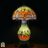 Verlichte Tiffany tafellamp Libellula Lumina ⌀30 op donkere achtergrond, koepelvormige kap met oranje en geel-groene panelen, libellenfries, bolvormige glas-in-lood voet met geel-groene en oranje panelen en gekleurde juwelen, bronzen leeuwenpootvoet, trekschakelaars