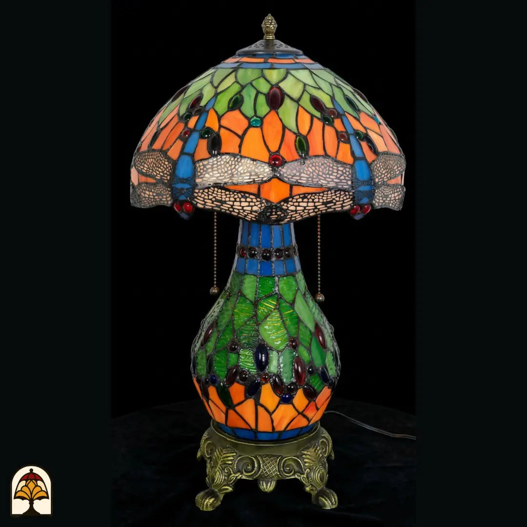 Onverlichte Tiffany tafellamp Libellula Lumina ⌀30 op donkere achtergrond, kap met groene en oranje panelen, blauwe libellenfries, gekleurde juwelen, groene glas-in-lood bolvoet, trekschakelaars, bronzen leeuwenpootvoet