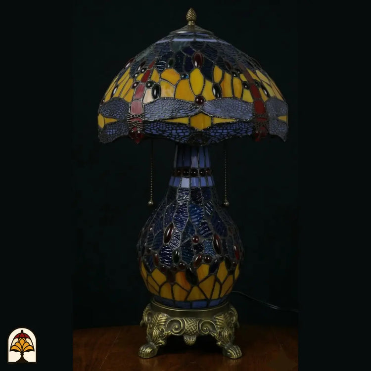 Onverlichte Tiffany tafellamp Libellula Azurea ⌀30 op donkere achtergrond, kap met gele en donkerblauwe panelen, paarse libellenfries, gekleurde juwelen, donkerblauwe glas-in-lood bolvoet met rode juwelen, bronzen leeuwenpootvoet