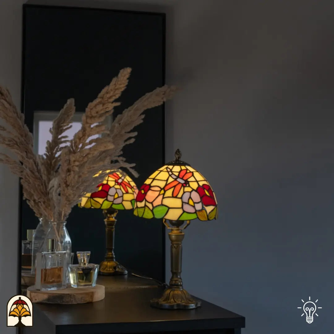 Lifestyle — verlichte Tiffany tafellamp op zwart dressoir voor grijze muur, spiegelreflectie, pampasgrassen en parfumflesjes ernaast