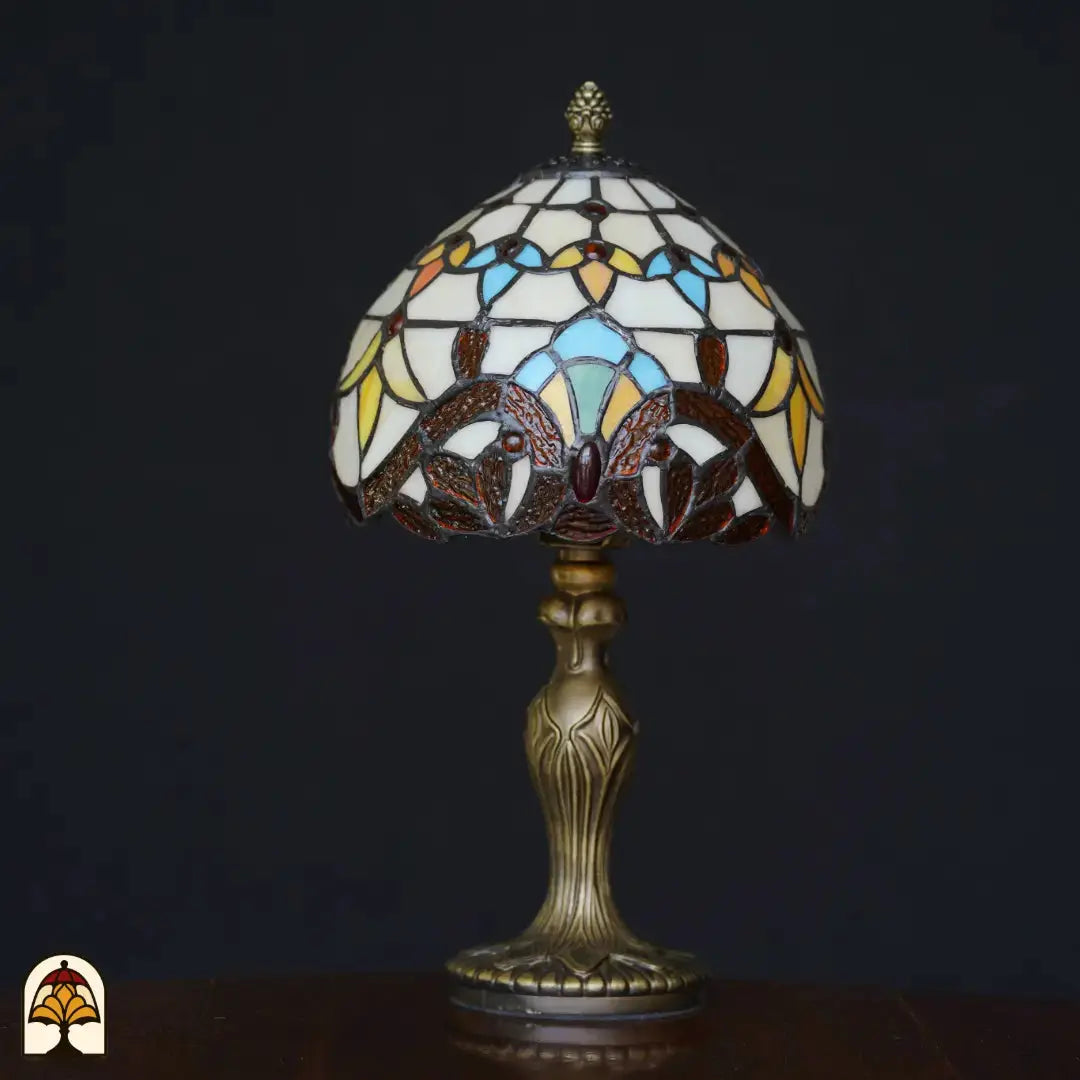 Onverlichte Tiffany tafellamp Baroque op zwarte achtergrond – koepelkap met crème, lichtblauw, geel en bruine barokke bladvormen, rode edelsteen