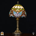 Verlichte Tiffany tafellamp Lucette ⌀20 cm op zwarte achtergrond – crème koepelkap met groene sierband, gouden libelle-/bladvormen, lichtblauwe en roze bloemblaadjes, rode edelstenen, bronzen kolomvoet