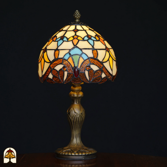 Klassieke Tiffany-stijl tafellamp die aan staat met sierlijke bronzen voet en glas-in-lood kap in geel-, blauw- en bruintinten, tegen een zwarte achtergrond.