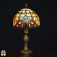 Klassieke Tiffany-stijl tafellamp die aan staat met sierlijke bronzen voet en glas-in-lood kap in geel-, blauw- en bruintinten, tegen een zwarte achtergrond.