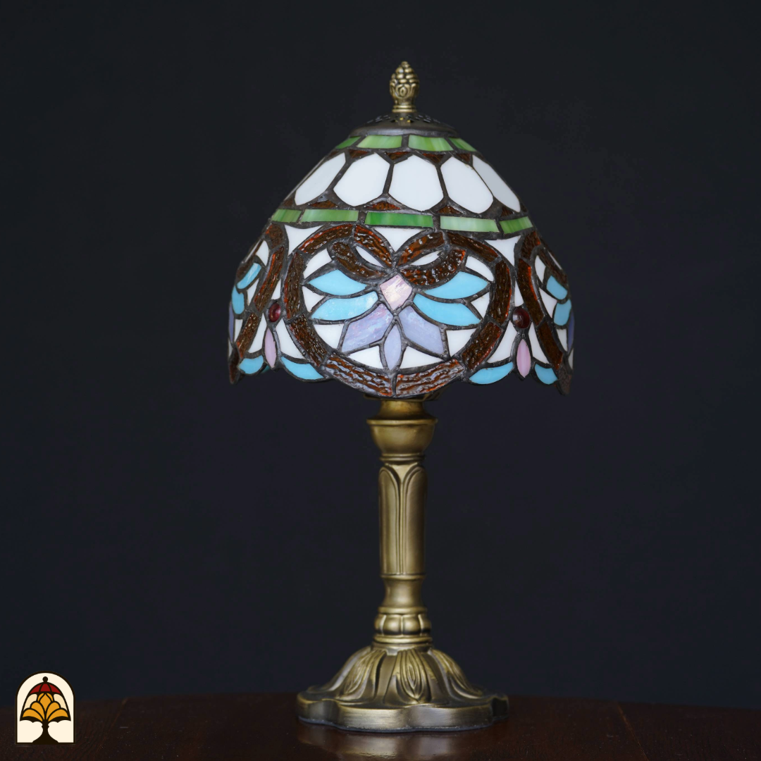 Tiffany-stijl tafellamp met glas-in-lood kap in pastelblauw, wit en groen, op een bronzen voet, geplaatst op een houten tafel tegen een zwarte achtergrond. De lamp staat uit.