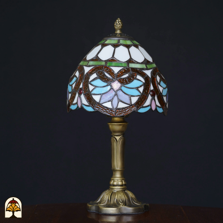 Tiffany-stijl tafellamp met glas-in-lood kap in pastelblauw, wit en groen, op een bronzen voet, geplaatst op een houten tafel tegen een zwarte achtergrond. De lamp staat uit.