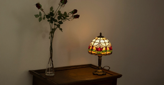 Brandende Tiffany-stijl tafellamp met rood tulpmotief en glas-in-lood kap, geplaatst op een houten kastje naast een vaas met gedroogde rode rozen, met schilderijen aan de muur erboven.
