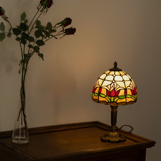 Brandende Tiffany-stijl tafellamp met rood tulpmotief en glas-in-lood kap, geplaatst op een houten kastje naast een vaas met gedroogde rode rozen, met schilderijen aan de muur erboven.
