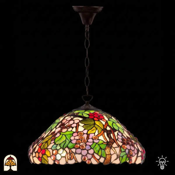Verlichte Tiffany hanglamp Sylva Berria aan bronzen ketting, koepelvormige kap met wijnrankmotieven, rode druiventrosjes en lila-roze druivenkorrels en groene bladeren in glas-in-lood, donkere achtergrond