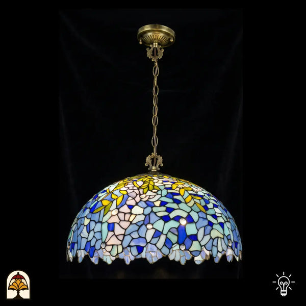 Verlichte Tiffany hanglamp Drizzle Bleu ⌀50 op donkere achtergrond, grote koepelvormige kap met blauwe en turquoise glas-in-lood bloemenmotieven, gele bladaccenten, geschulpte onderrand, bronzen ketting en plafondrozet