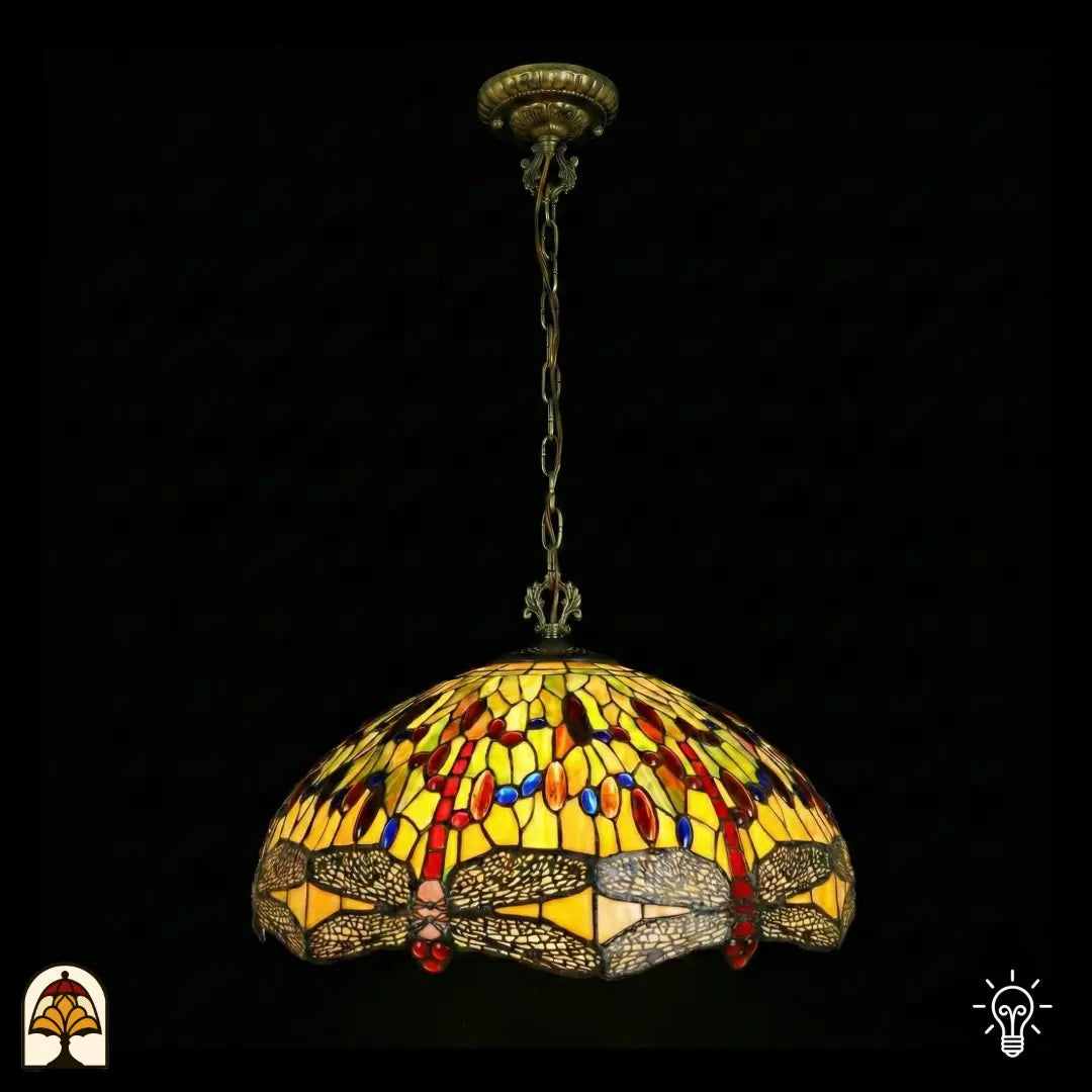 Onverlichte Tiffany hanglamp Libellula Aurelia ⌀50 op donkere achtergrond, grote koepelvormige kap met gele glas-in-lood panelen, libellenfries onderaan, rode en blauwe juwelen, bronzen ketting en plafondrozet
