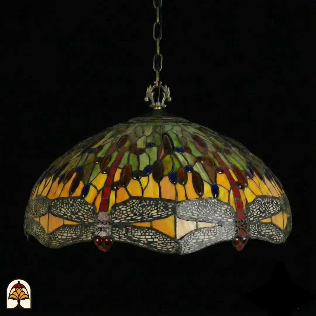 Onverlichte Tiffany hanglamp Libellula Aurelia ⌀50 op donkere achtergrond, kap met gele en groene panelen, grijze libellenfries met rode juwelen langs rand, bronzen ketting