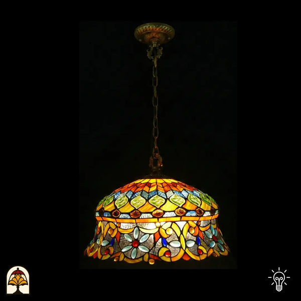 Tiffany lamp hanglamp verlicht, koepelvormige kap met amberkleurige bovenzone met groene diamantvormige glaskralen en blauw mozaïekglas, onderzone met gele arabesken en lichtblauwe bloemkralen op transparant glas, gekartelde rand, opgehangen aan bronzen ketting