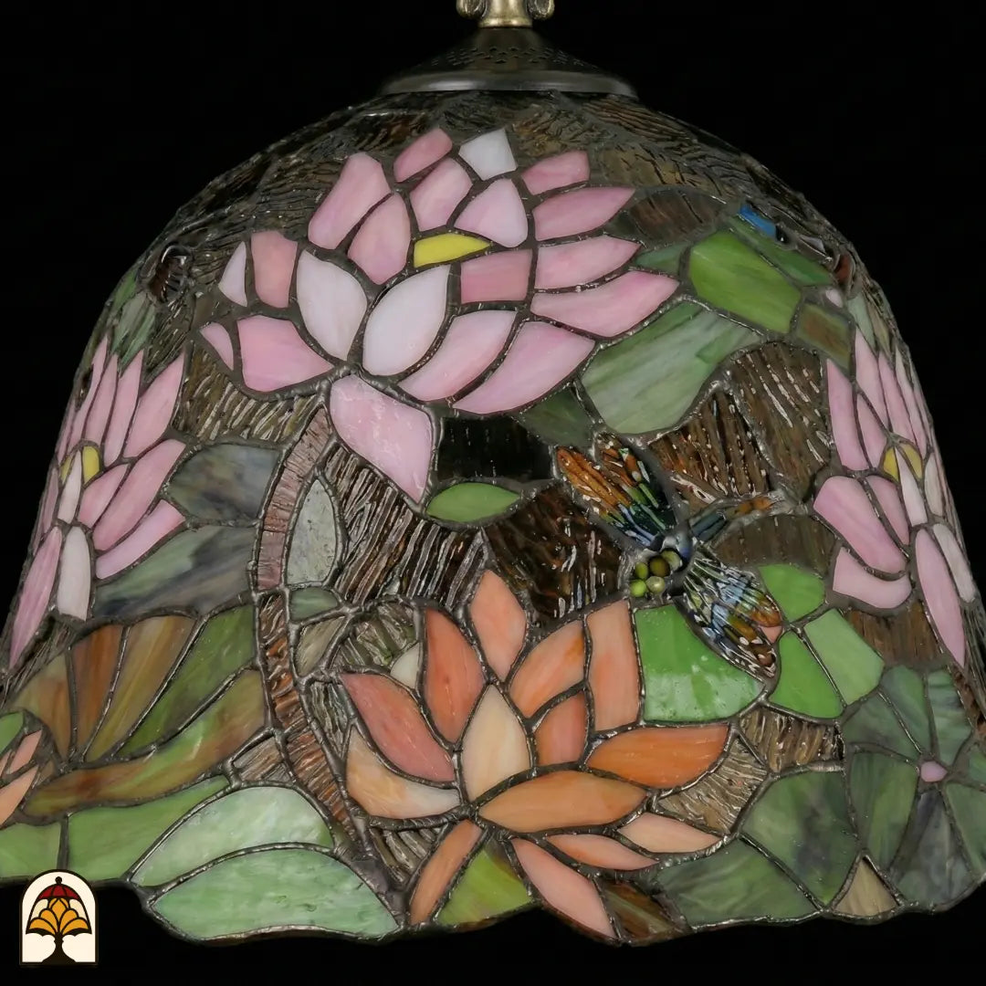 Detailopname van tiffany lamp klokvormige kap onverlicht met roze lotusbloem centraal, oranje lotusbloemen onderaan, groene waterleliebladen en een bruinoranje libelle met blauwe details