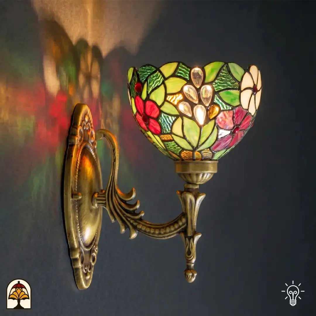 Tiffany lamp wandlamp verlicht aan muur, komvormige glas-in-lood kap met rode en roze bloemen, groene bladeren en transparante glaskralen; sierlijke bronzen muurbeugel met ovale wandplaat en acanthusrolwerk; kleurrijke lichtreflecties op de muur