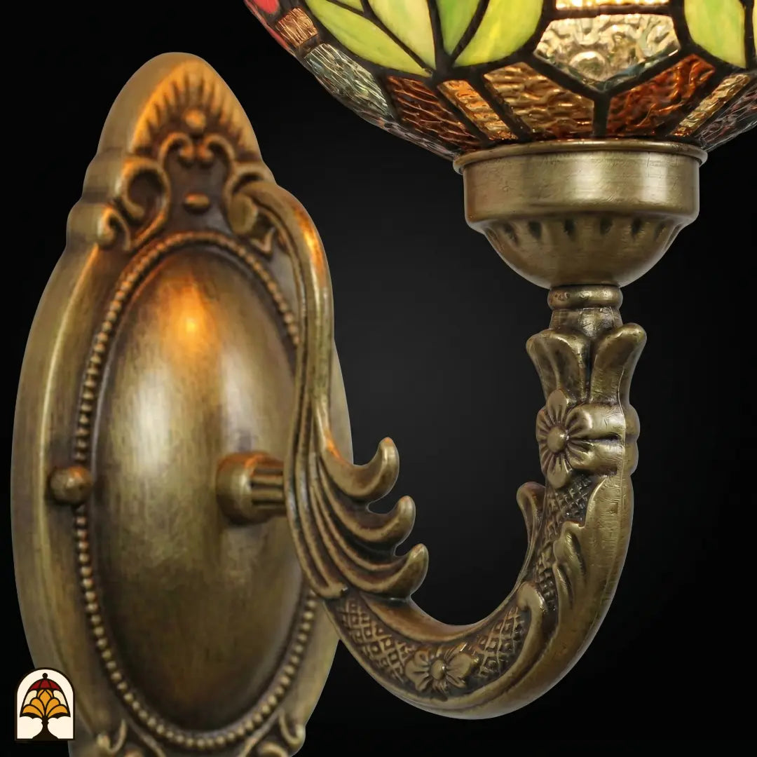 Close-up van bronzen wandbeugel met ovale reliëfplaat en gebogen arm, bovenkant van de florale glas-in-lood kap met groen en amber zichtbaar