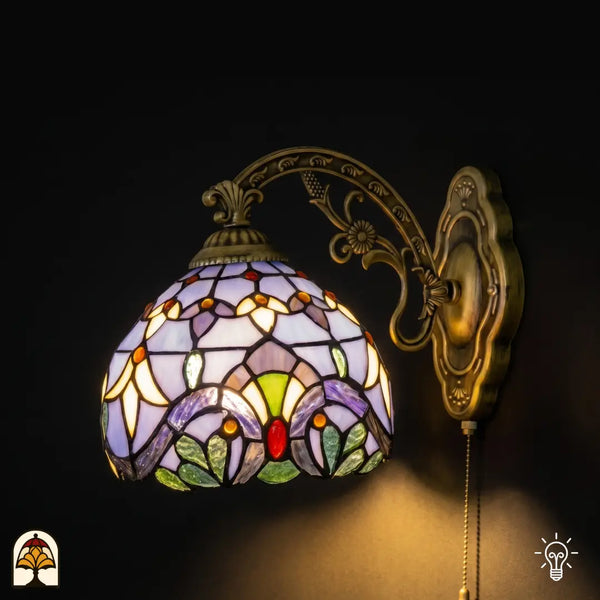 Verlichte Tiffany wandlamp Royal Mosaic met lavendelblauwe glas-in-lood kap, crèmegele bloemmotieven en kleurrijke juwelen op bronzen sierlijke arm
