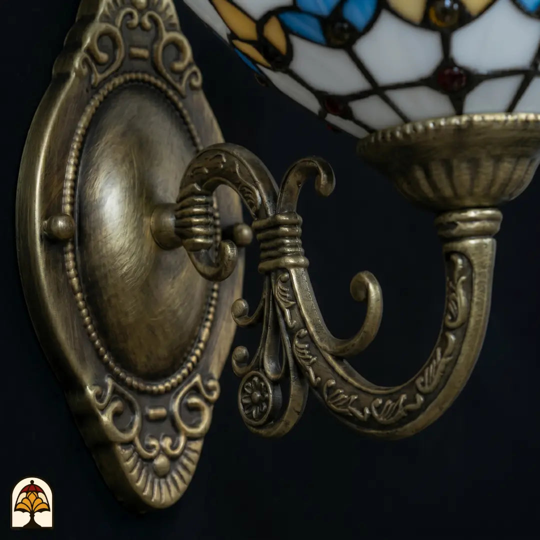 Close-up van de bronzen wandbeugel met ovale reliëfplaat en gebogen arm, goudgele glas-in-lood kap van de Tiffany wandlamp gedeeltelijk zichtbaar