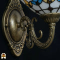 Close-up van de bronzen wandbeugel met ovale reliëfplaat en gebogen arm, goudgele glas-in-lood kap van de Tiffany wandlamp gedeeltelijk zichtbaar