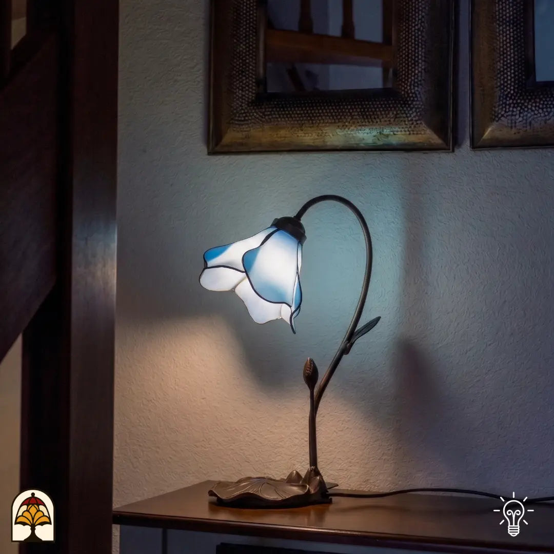 Een sfeervolle Tiffany tafellamp in de vorm van een blauw-witte bloemkelk, rustend op een sierlijke bronskleurige voet met bladmotieven. De lamp staat op een houten kastje tegen een witte muur, met op de achtergrond twee decoratieve spiegels en een houten trap. De lamp verspreidt een zacht, blauwachtig licht in een sfeervol verlichte kamer.