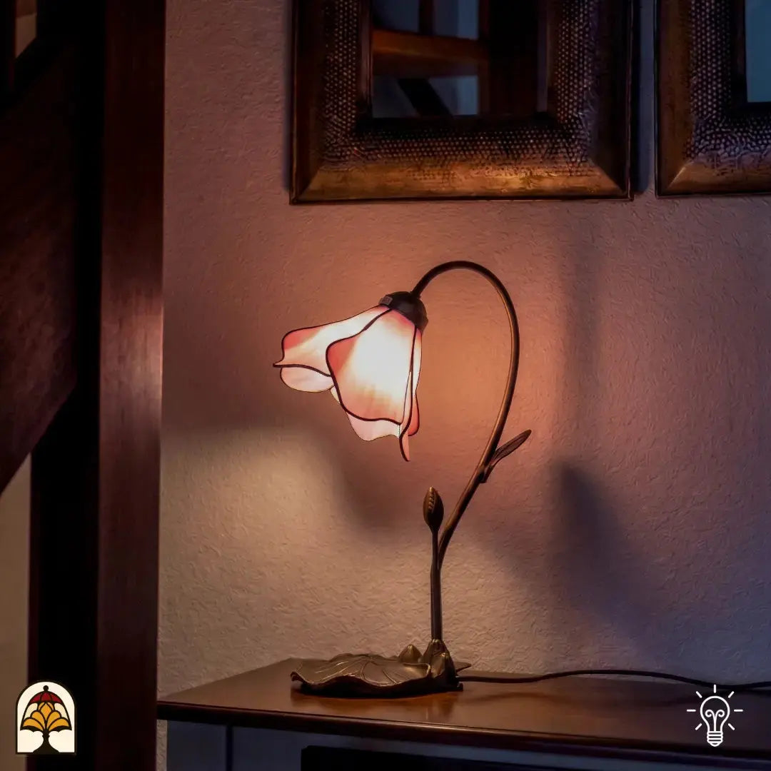 Een sfeervolle Tiffany tafellamp in de vorm van een zachtroze bloemkelk, gemonteerd op een sierlijke bronskleurige voet met een knopje en bladmotief. De lamp staat op een houten sidetable en verspreidt een warm, roze licht tegen een witte wand. Op de achtergrond hangen twee vierkante, gedecoreerde spiegels naast een houten trappartij.