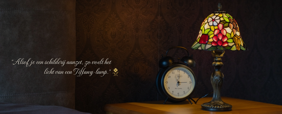 Handgemaakte Tiffany tafellamp met kleurrijke kap in druiven- en bloemmotief, geplaatst op een nachtkastje naast een klassieke wekker. Sfeervolle lamp die warmte en nostalgie in de slaapkamer brengt.