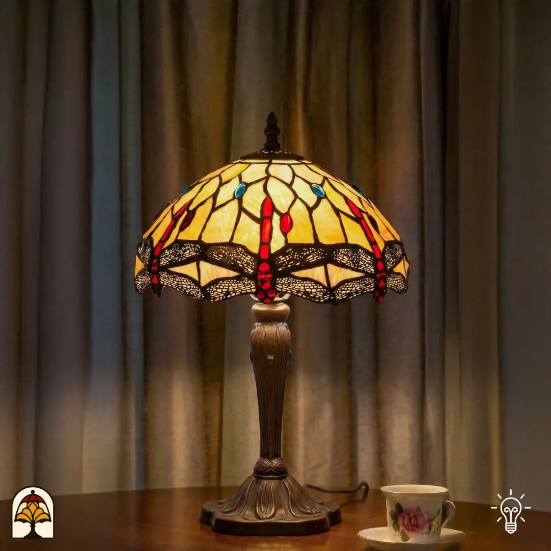 Verlichte Tiffany tafellamp Libelle Elegance op houten tafel voor gordijnen, geel-amber kap met rode accenten en libellenmotieven, theekopje ernaast