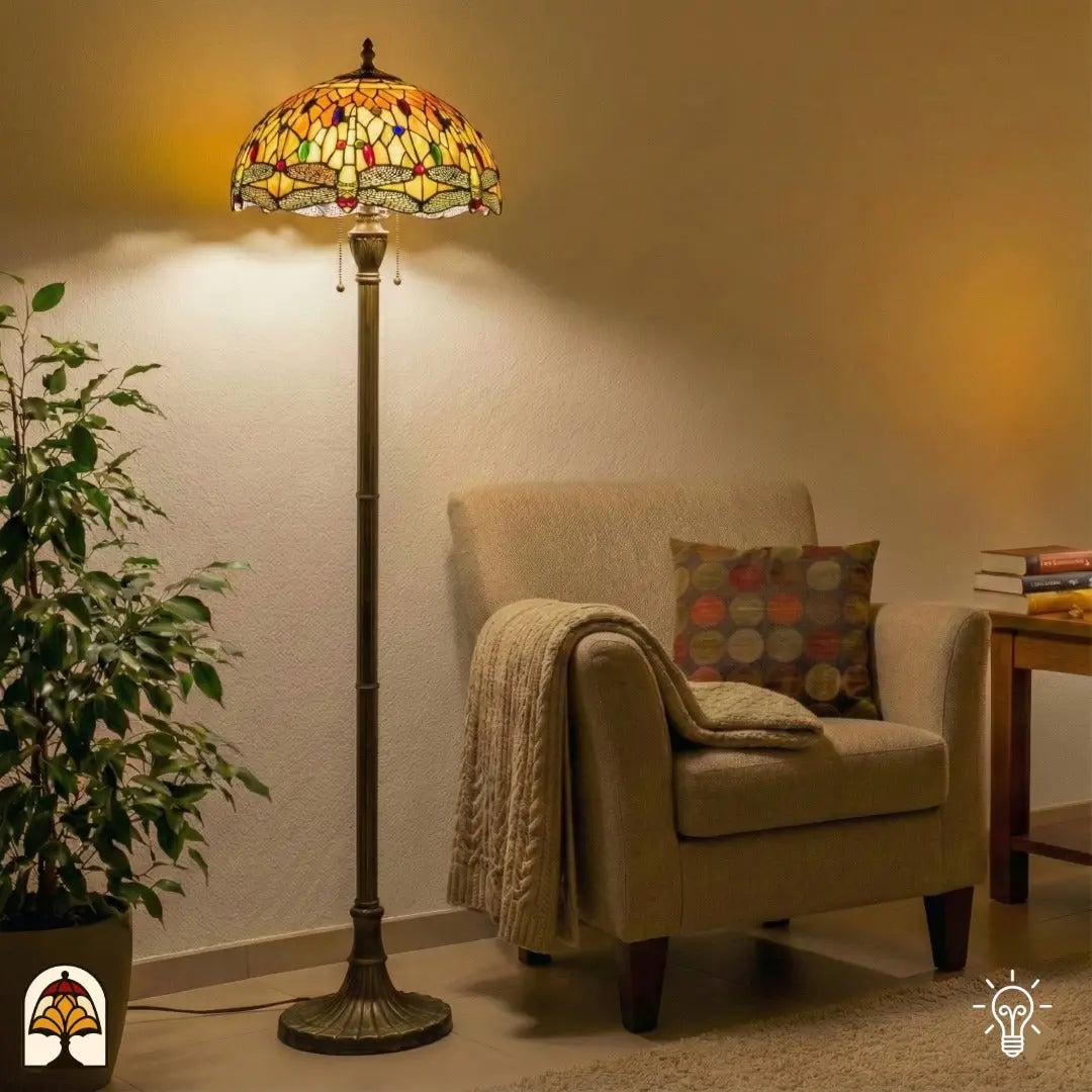Tiffany vloerlamp Libellula Aurea verlicht in woonkamer — naast beige fauteuil met deken en kussen, groene plant links, bijzettafel met boeken rechts
