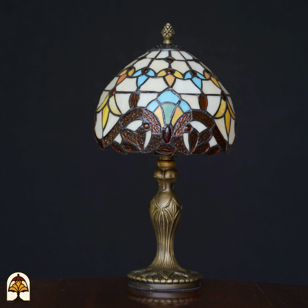 Tiffany-stijl tafellamp die uit staat met glas-in-lood kap in geel-, blauw- en bruintinten en een sierlijke bronzen voet, geplaatst op een houten tafel tegen een zwarte achtergrond.