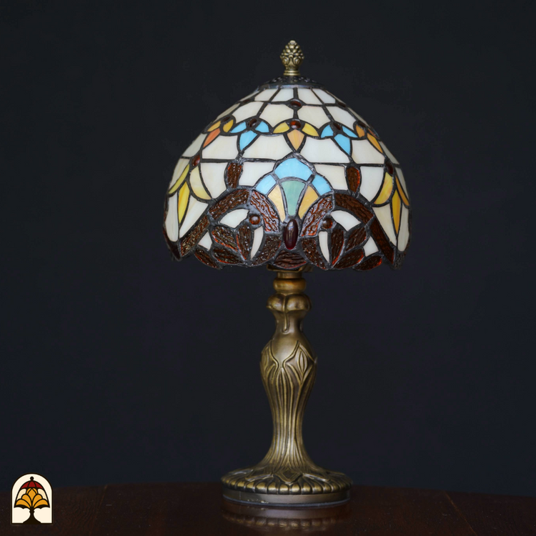 Tiffany-stijl tafellamp die uit staat met glas-in-lood kap in geel-, blauw- en bruintinten en een sierlijke bronzen voet, geplaatst op een houten tafel tegen een zwarte achtergrond.