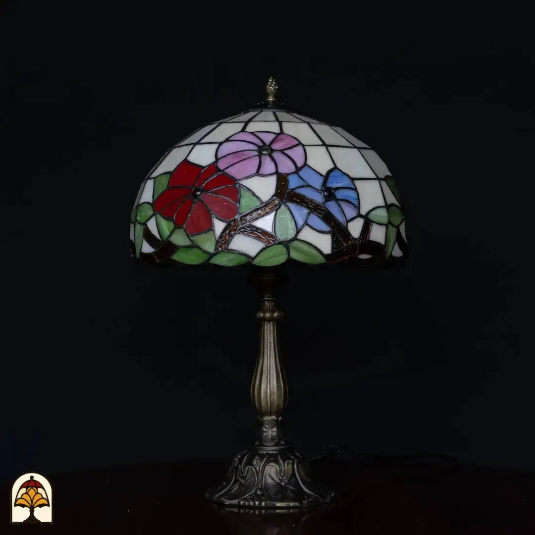 Tiffany tafellamp Aurora Bloom onverlicht op zwarte achtergrond — witte kap met roze, rode en blauwe bloemen, gouden takken, groene bladeren, bronzen kolomvoet