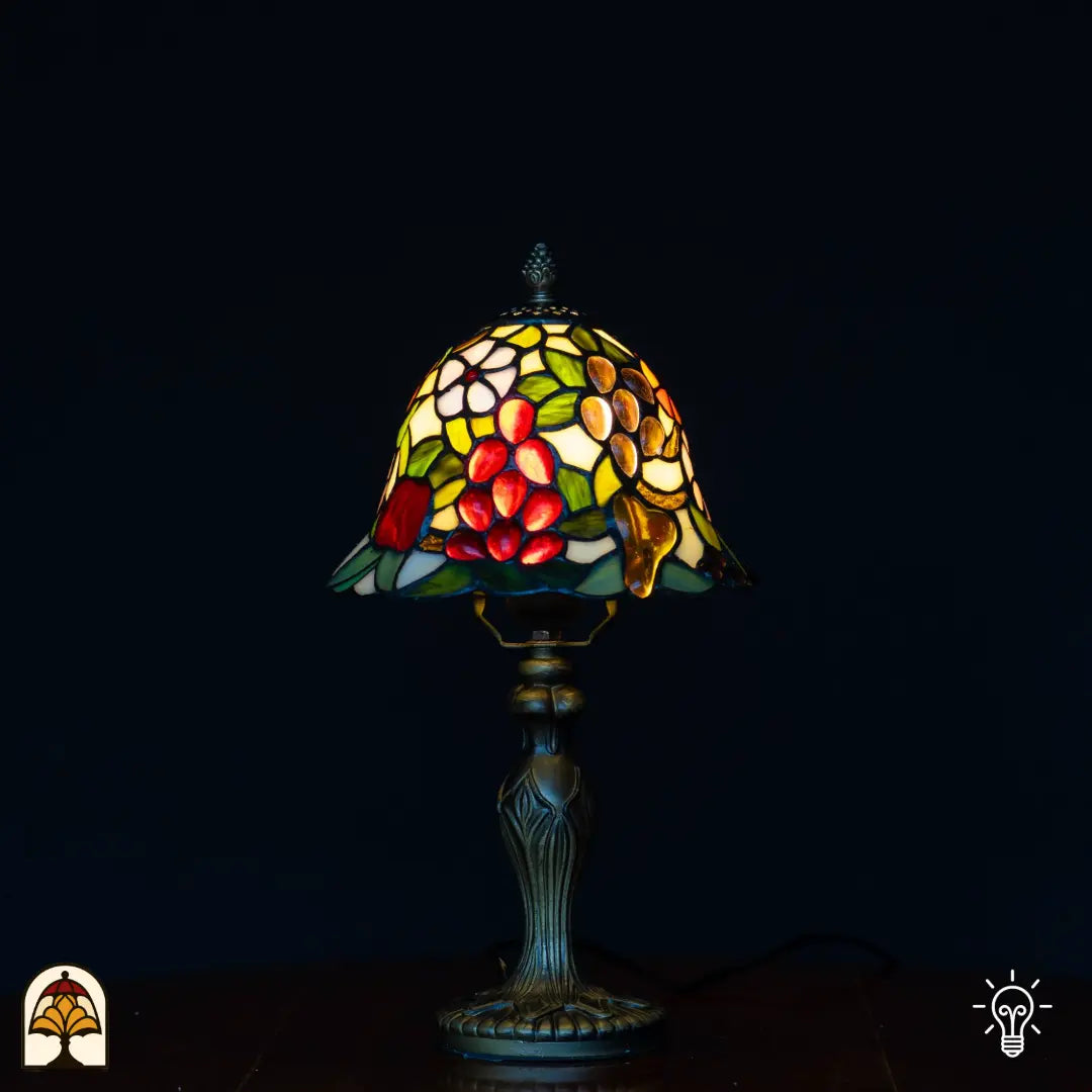 Verlichte Tiffany tafellamp met klokvormige kap met rode druiven, amber/gouden peervormige vruchten, witte bloemen en groene bladeren op gele achtergrond, klassieke kandelaarvoet, zwarte achtergrond
