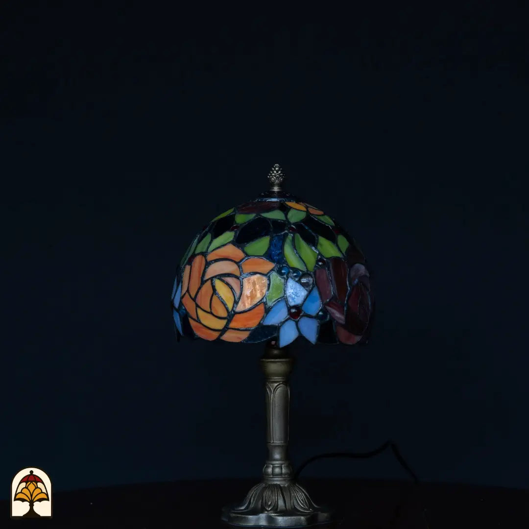 Tiffany tafellamp Fiore onverlicht op donkere achtergrond — kap met oranje rozen, groene bladeren, blauwe bloemen en paarse roos, bronzen kolomvoet