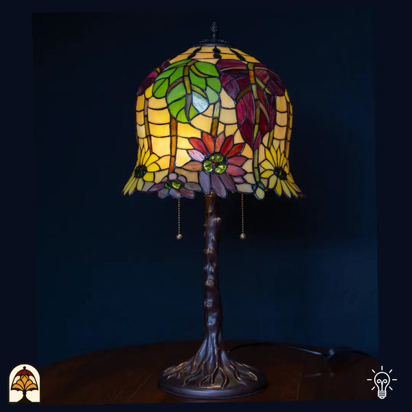 Verlichte tafellamp met hoge klokvormige amber/gele Tiffany kap met rode en paarse bloemen en groene bladeren, boomstam-voet met trekschakelaars, donkerblauwe achtergrond