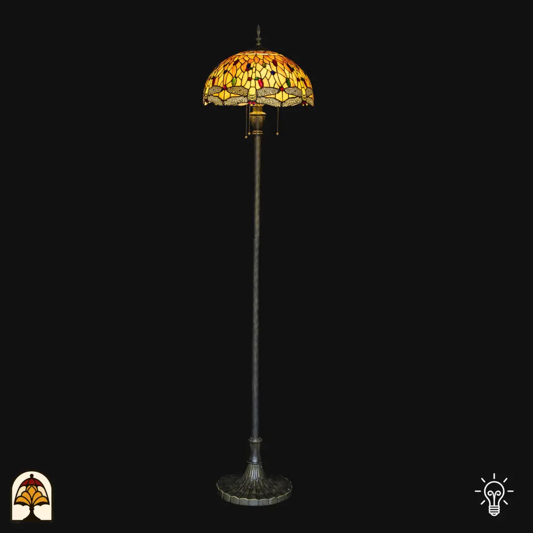 Tiffany vloerlamp Libellula Aurea verlicht op zwarte achtergrond — amber/gele koepelkap met libellenrand, lange paal met brede bronzen voet, trekschakelaars